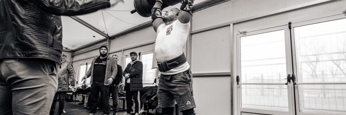 A strongman lifts a barbell overhead during an indoor gym event, showcasing strength and determination.